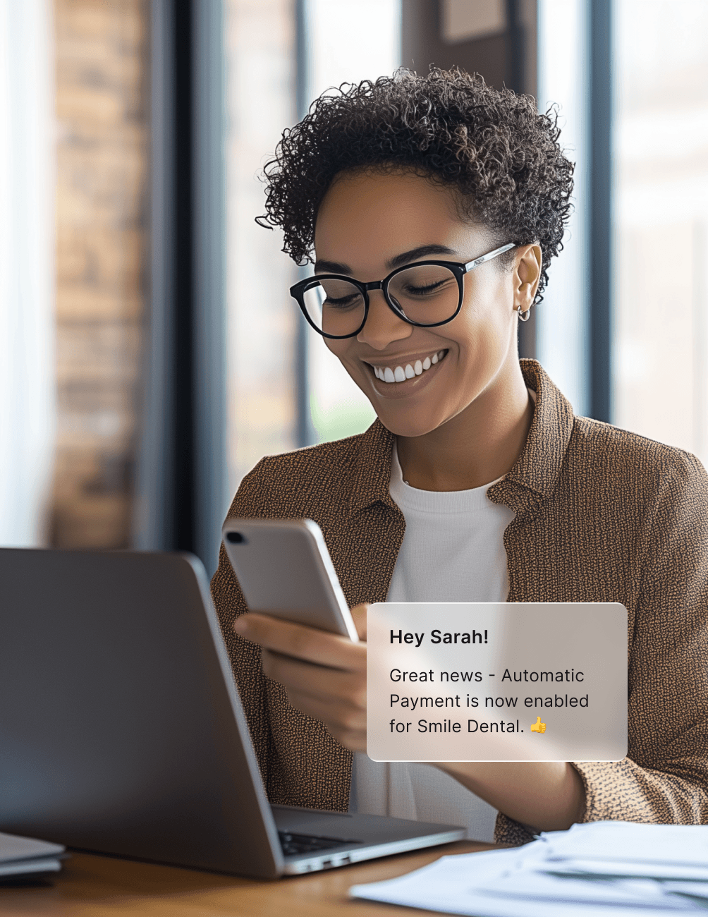 A woman smiles at her phone while sitting at a desk with a laptop. A message on the screen says automatic payment is enabled for Smile Dental.