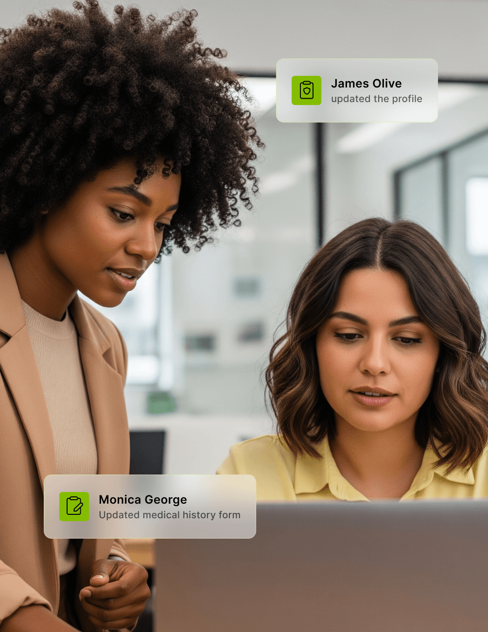 Two women at a desk, one standing and one sitting, looking at a laptop. Notifications about profile updates are displayed.