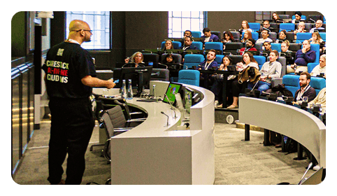 A speaker presents to an attentive audience in a modern lecture hall with tiered seating and multiple screens.